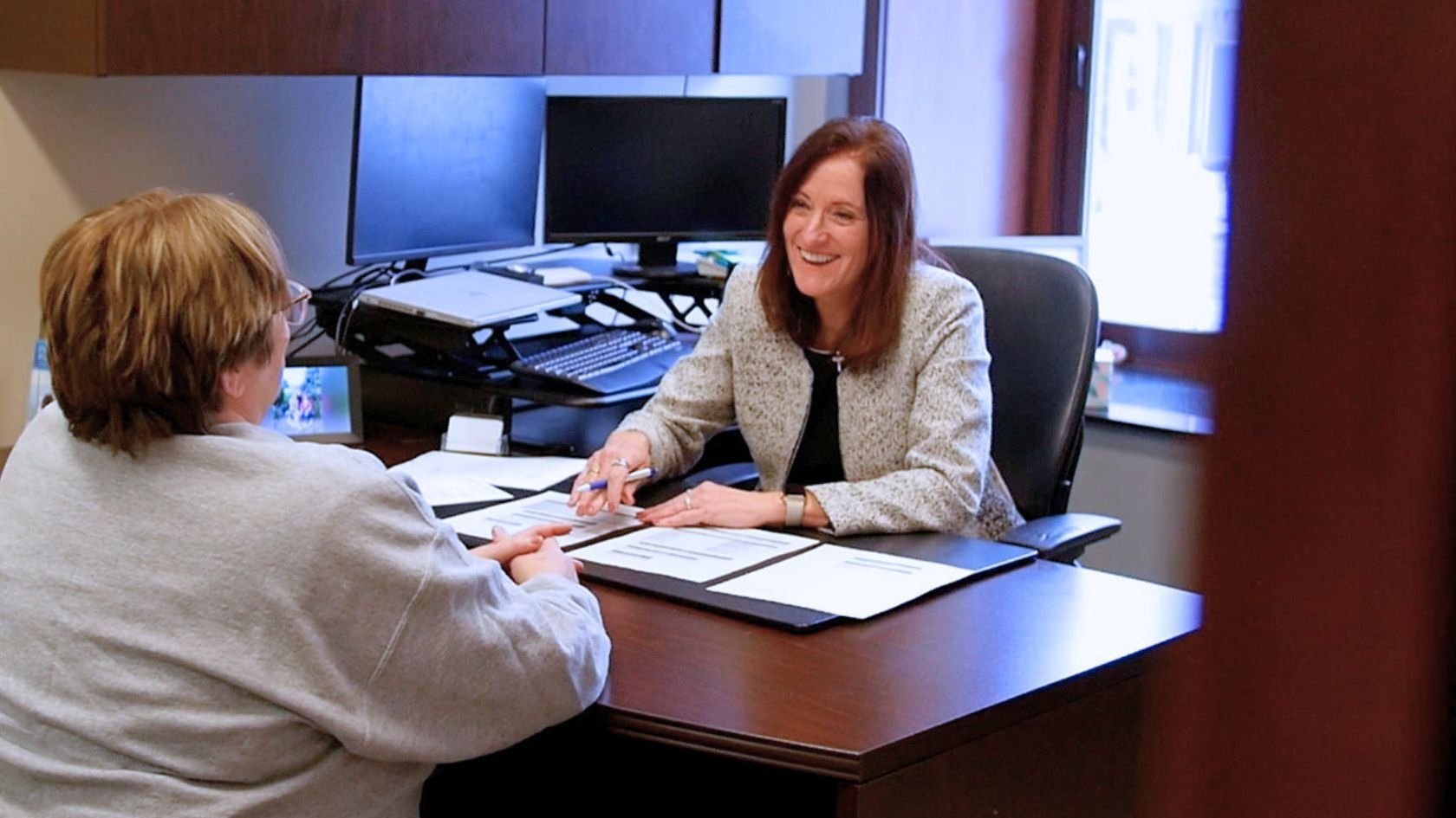 A Heartland Bank Relationship Manager smiles while meeting with a customer across a desk, discussing personal loan options in an office setting with a computer and files in the background.