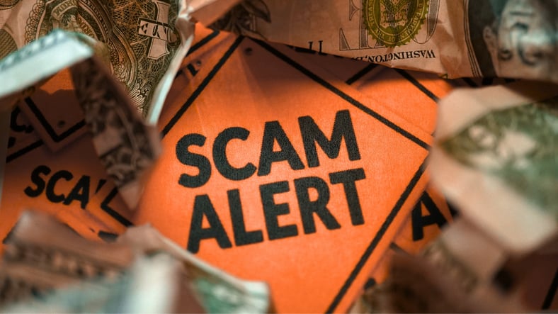 An orange warning sign reading “Scam Alert” is surrounded by crumpled U.S. dollar bills, symbolizing financial fraud and the risk of monetary loss from scams.